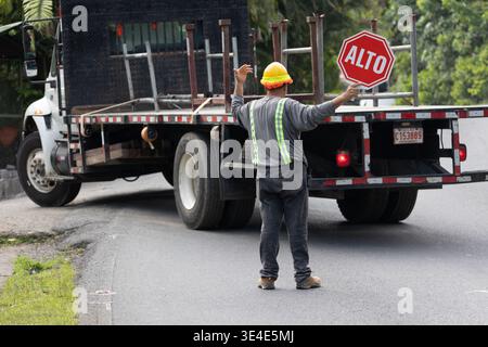 Manuel Antonio, Costa Rica - 9. August 2025: Eine Baustelle mit Schutzhelm hält ein „ALTO“-STOPPSCHILD hoch, während ein Lkw auf einer Straße rückwärts fährt Stockfoto