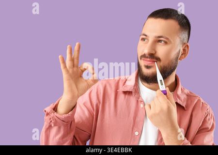 Hübscher junger Mann mit Thermometer, das OK-Geste auf Flieder-Hintergrund zeigt Stockfoto