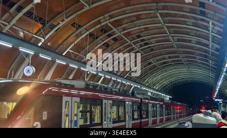Moderner Bahnsteig bei Nacht mit rot-weißem Zug, der unter dem beleuchteten geschwungenen Dach hielt, Pendler warteten. Stockfoto
