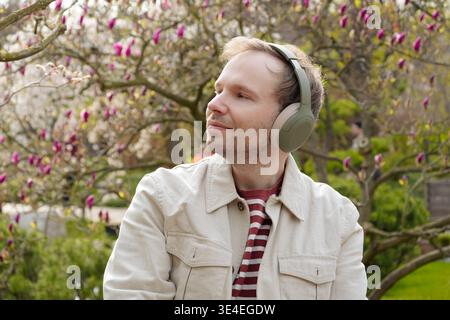 Ein lächelnder Mann mit Kopfhörern entspannt sich in der Natur zwischen blühenden Magnolien in einem japanischen Park und genießt eine digitale Entgiftung. Stockfoto