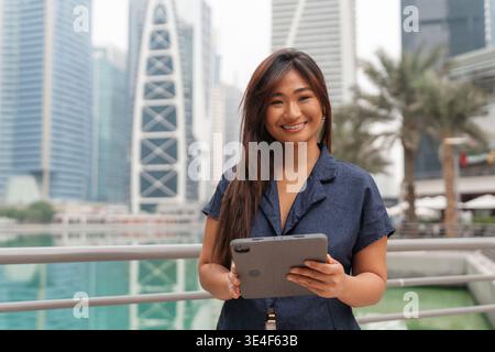 Geschäftsfrau steht im Finanzviertel von Dubai, hält ein Tablet und lächelt und zeigt modernes Unternehmertum in der Golfregion. Stockfoto