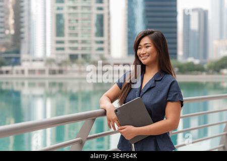 Eine Geschäftsfrau steht in Dubai am Wasser und lächelt selbstbewusst, während sie ein Tablet hält. Sie steht für Erfolg im Finanzwesen. Stockfoto