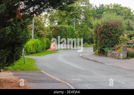 Eine ruhige asphaltierte Straße führt durch ein friedliches englisches Dorf, das von üppigen grünen Hecken, Steinmauern und alten Bäumen umgeben ist. Stockfoto