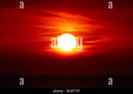 Ein lebhafter Sonnenuntergang über dem Ozean erfüllt den Himmel mit intensiven Rot- und Orangentönen, während die helle Sonne über dem Horizont schwebt. Stockfoto