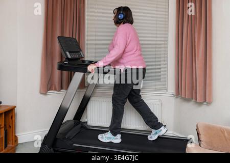 Weite Aufnahme der Frau, die auf dem Laufband im Wohnraum läuft, zeigt das Heim-Gym-Setup, Indoor-Training und gesunde Lebensstil-Routine Stockfoto