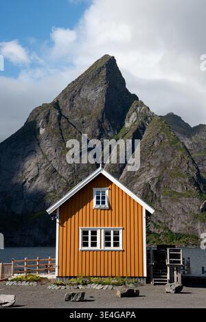 Gelbes Holzhaus liegt in der Nähe des Wassers mit einem hohen Berg im Hintergrund an einem klaren Tag. Lofoten-Inseln Norwegen Stockfoto