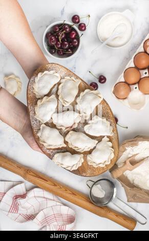 Essen, bereiten Sie köstliche süße Teigtaschen mit Kirschen zu. Traditionelles russisches und ukrainisches Dessert. Vareniki und Zutaten auf dem Marmorhintergrund -f Stockfoto