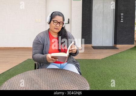 Frau, die sich auf ein Buch konzentriert, während sie Kopfhörer trägt und an einem Tisch sitzt Stockfoto