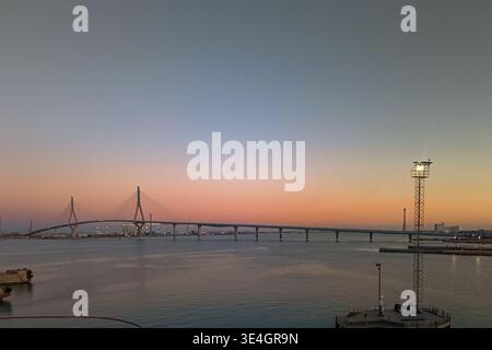Kabelbrücke über ruhiges Wasser bei Sonnenuntergang mit weichem Pastellhimmel, Küsteninfrastruktur und ruhiger maritimer Landschaft. Stockfoto