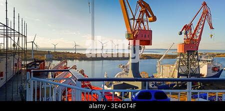 Blick von einem Schiffsdeck auf Hafenkräne und Offshore-Windturbinen in der Ferne, industrielle maritime Szene mit Infrastruktur für erneuerbare Energien. Stockfoto