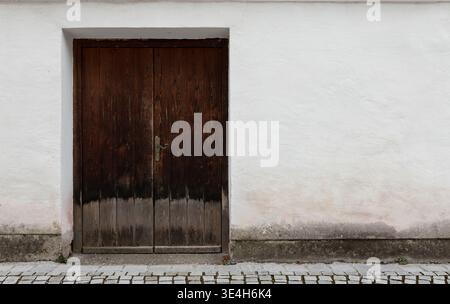 Verwitterte dunkle Holztür in einer weißen Putzwand mit Kopfsteinpflastervordergrund und offenem Wandraum, ideal für Architektur und Design. Stockfoto