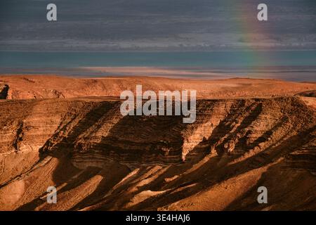 Heller Regenbogen über dramatischer Wüstenlandschaft mit Sturmwolken und ruhigem Meer im Hintergrund. Stimmungsvolle Naturkulisse mit farbenfrohen Lichtkontra Stockfoto