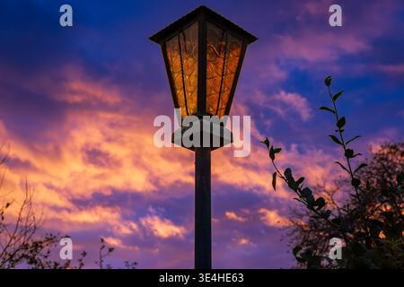 Nahaufnahme einer alten Straßenlaterne, die in der Abenddämmerung vor einem farbenfrohen Sonnenuntergangshimmel mit dramatischen Wolken leuchtet. Stimmungsvolle Abendlichtszene. Stockfoto