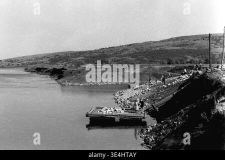 Nikolajewka, 1957. Slawjanskaja GRES. Vorbereitungsarbeiten in der Nähe eines Salzsees (wahrscheinlich Repnoye) für den massiven Kraftwerksbau SlavGRES. Mehrere Arbeiter werden am Ufer stehend und arbeiten gesehen, während ein großes Holzfloß im ruhigen Wasser mit schweren Steinmaterialien schwimmt. Der ferne Hintergrund zeigt eine natürliche Landschaft mit sanften Hügeln unter klarem Himmel. Dieses Vintage-Schwarzweißfoto erfasst die frühen Vorbereitungsphasen eines großen Industrieprojekts, das das friedliche Leben und die Entwicklung der Vorkriegszeit im friedlichen Donbass widerspiegelt. Stockfoto