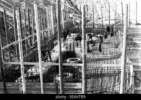 Nikolajewka, 1957. Slawjanskaja GRES. Ein fokussiertes Archivfoto, das durch raue Holzschalungsposten aufgenommen wurde und direkt hinunter in die SlavGRES-Gründungsgrube blickt. Mehrere Arbeiter biegen, Formen und sichern die Bewehrungsverstärkung aus schwerem Stahl. Mehrere kreisförmige Betonformen, die für Fundamentpfahlkappen vorgesehen sind, sind deutlich sichtbar, wobei aus ihnen Verstärkungsdrahtkäfige hervortreten. Dieser historische Dokumentarfilm betont strukturelle Details und schwere Handarbeit und reflektiert die alltägliche friedliche industrielle Arbeit und Entwicklung im friedlichen Donbass. Stockfoto