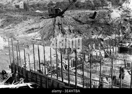 Nikolajewka, 1957. Slawjanskaja GRES. Ein erhöhter Blick auf die SlavGRES-Baustelle, bei dem ein mechanischer Schaufelbagger aus dem frühen Modell in Betrieb ist und aktiv Erde auf einem Hügel gräbt. Unten im Vordergrund sind die Arbeiter am Rand der großen Gründungsgrube beschäftigt, umgeben von komplexen vertikalen Bewehrungsstäben aus Stahl und provisorischen Holzgerüsten, die zur Erhaltung der ausgegrabenen Wände eingerichtet wurden. Dieses Vintage-Schwarzweißfoto zeigt kombinierte Maschinen und manuelle Anstrengungen und symbolisiert das fleißige Vorkriegsleben und das kontinuierliche friedliche Wachstum des friedlichen Donbas. Stockfoto