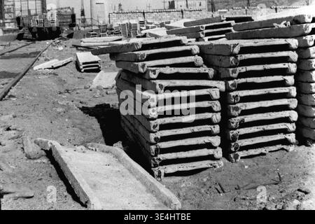 Nikolajewka, 1957. Slawjanskaja GRES. Ein Nahaufnahme-Archivfoto, das sich speziell auf schwere Stapel von gerippten Betonfertigteilen konzentriert, die sich auf einem Lagerplatz für den Bau des Kraftwerks SlavGRES befinden. Im fernen Hintergrund sind ein Eisenbahngleis und möglicherweise ein Bahnsteig zu sehen, was die Versorgungslogistik auf der Schiene angibt. Die gerippte Struktur der Betonformen ist hier ein sehr markantes Merkmal. Diese Vintage-Aufnahme veranschaulicht die notwendige Materiallieferkette und erfasst friedliche Industriearbeiten aus der Vorkriegszeit, Logistik und Wachstum im friedlichen Donbass. Stockfoto