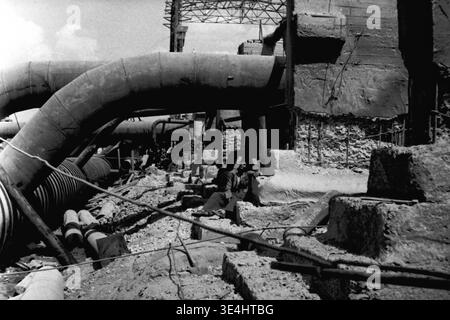 Nikolajewka, 1957. Slawjanskaja GRES. Ein beeindruckendes Archivfoto, das die Installation von gebogenen Stahlrohren mit großem Durchmesser für das Kraftwerk SlavGRES feststellt. Diese massiven Industrierohre werden auf provisorischen Stützen an der Basis des Grabens verlegt. Ein einziger Arbeiter ist deutlich sichtbar, der ruhig im tiefen Schatten unter einem der Rohre sitzt und ruht. Dieses Vintage-Bild zeigt den beeindruckenden technischen Maßstab und die menschliche Präsenz, die den friedlichen Alltag und die hart arbeitende industrielle Entwicklung im friedlichen Donbass vor dem Krieg widerspiegelt. Stockfoto