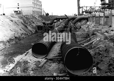 Nikolajewka, 1957. Slavyanskaya GRES Ein eindrucksvolles perspektivisches Foto, das gerade entlang eines neu installierten Rohrleitungsgrabens mit großem Durchmesser für die SlavGRES-Konstruktion blickt. Ein Einzelarbeiter steht direkt neben einem der massiven Wellstahlrohre, die der enormen Ingenieurstruktur eine perfekte Größe verleihen. Im fernen Hintergrund ist das aufsteigende Stahlgerüst des Hauptgebäudes mit einem Kran sichtbar. Dieses Vintage-Bild spiegelt das große Ausmaß der industriellen Entwicklung wider und spiegelt fleißiges friedliches Vorkriegsleben und Wachstum im friedlichen Donbass wider. Stockfoto