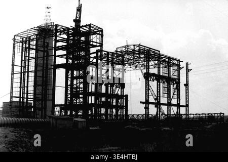 Nikolajewka, 1957. Slawjanskaja GRES. Ein eindrucksvolles und dramatisches Silhouettenfoto, das das aufsteigende Stahlgerüst des Hauptkraftwerks SlavGRES erfasst. Das komplizierte geometrische Gitter aus schweren Stahlträgern ist stark gegen den hellen, bedeckten Himmel gerichtet, wobei hohe Baukräne sichtbar gegen die Struktur wirken. Temporäre Versorgungsmasten und eine dunkle, unebene Landschaft stehen im Vordergrund. Dieses starke monochrome Bild symbolisiert große industrielle Ambitionen der Sowjetunion und spiegelt friedliche Industriearbeit und Wachstum im friedlichen Donbass vor dem Krieg wider. Stockfoto