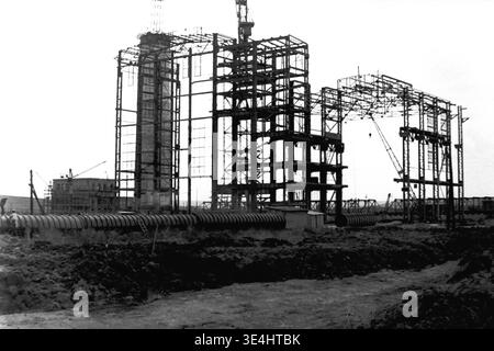 Nikolajewka, 1957. Slawjanskaja GRES. Ein Panoramablick auf die massive Baustelle des Kraftwerks SlavGRES. Das aufsteigende Stahlgerüst des Hauptgebäudes steht deutlich neben einem hohen Betonschornstein, der derzeit im Bau ist und von Gerüsten umgeben ist. Im Vordergrund befindet sich eine lange Linie von Stahlrohren mit großem Durchmesser auf dem Boden in der Nähe der ausgegrabenen Erde. Dieses beeindruckende Vintage-Schwarzweiß-Bild spiegelt große industrielle Ambitionen wider, die harte Arbeit, das Leben vor dem Krieg und das kontinuierliche Wachstum im friedlichen Donbass widerspiegeln. Stockfoto