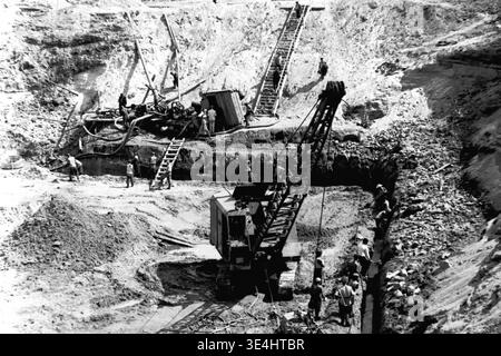 Nikolajewka, 1957. Tiefgründungsgrube des Kraftwerks Slavyanskaya GRES, aufgenommen in einem hochdynamischen Foto. Unten arbeitet ein schwerer Raupenkran, umgeben von Arbeitern, die manuell Gräben ausheben. Eine lange, steile Holztreppe bietet einen unentbehrlichen Zugang über den steilen Sandhang. Auf einer mittleren Plattform verwalten die Arbeiter schwere Pumpanlagen und dicke Schläuche. Dieses beeindruckende dokumentarische Bild zeigt komplexe, koordinierte Erdwerke, die das fleißige Vorkriegsleben und die schnelle industrielle Entwicklung im friedlichen Donbass widerspiegeln. Stockfoto