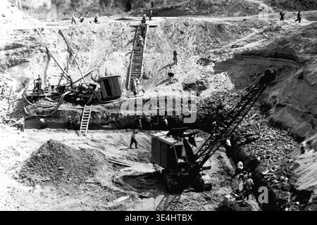 Nikolajewka, 1957. Slavyanskaya GRES Kraftwerk aktive Grundgrubenaushub aus einem anderen faszinierenden Winkel gesehen. An der Basis arbeitet ein großer mechanischer Raupenkran, während zahlreiche Arbeiter intensive manuelle Erdbewegungsarbeiten in der Nähe ausführen. Mehrere Arbeiter sind deutlich beim Aufstieg der steilen provisorischen Holztreppe zu sehen, die direkt in den ausgegrabenen Hang gebaut wurde. Dieser Vintage-monochrome Schnappschuss zeigt die Mischung aus schweren Maschinen und menschlichem Einsatz und demonstriert ehrgeiziges Vorkriegsleben und große industrielle Expansion im friedlichen Donbass. Stockfoto