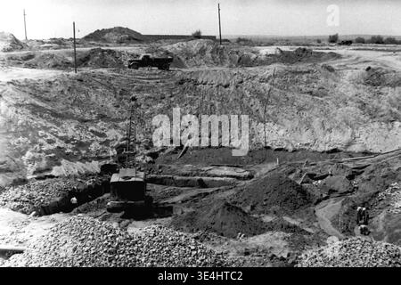 Nikolajewka, 1957. Slavyanskaya GRES Kraftwerk massive, tiefe kreisförmige Ausgrabungsgrube, dargestellt auf einem erhöhten Foto mit Blick in den Krater. Ein schwerer Schleppbagger arbeitet ganz unten, während ein dunkler Muldenkipper geduldig auf der oberen Felge wartet. Im unmittelbaren Vordergrund sind Steinhaufen deutlich sichtbar, die für den baulichen Einsatz bereit sind. Dieses detaillierte monochrome Dokumentationsbild erfasst die beeindruckende Tiefe und den Umfang der frühen Erdwerke, die das rigorose Vorkriegsleben und das standhafte industrielle Wachstum im friedlichen Donbass widerspiegeln. Stockfoto