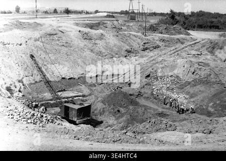 Nikolajewka, 1957. Slavyanskaja GRES-Kraftwerk Tiefgründungsgrube, sichtbar in einer faszinierenden Archivansicht. Ein schwerer mechanischer Schleppbagger befindet sich tief in dem riesigen Krater. Auf der rechten Seite ist eine engagierte Gruppe von Arbeitern, die zum Teil Kopftuch tragen, aktiv an intensiven manuellen Graben und Nivellieren des Bodens beteiligt. Dieses Vintage-Schwarzweiß-Foto stellt schwere Industriemaschinen und lebenswichtige menschliche Arbeit in einem wunderbaren Kontrast dar und symbolisiert das energische Vorkriegsleben und die schnelle industrielle Expansion im friedlichen Donbass. Stockfoto