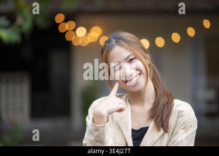 Glückliches, verspieltes Porträt junger asiatischer Frau mit einem hellen Lächeln, das mit dem Finger auf ihre Wange zeigt und mit einem niedlichen Zwinkern fröhliche Emotionen gegen die schöne zeigt Stockfoto