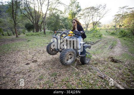 Glückliche junge Frau, die im Freien ein extremes ATV-Quad fährt und einen Wheelie-Trick in einem wunderschönen, sonnigen grünen Naturwald mit aufgeregtem emotionalen Ausdruck macht Stockfoto