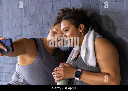 Afroamerikanische Freundinnen in Tanks, die sich an die Wand des Fitnessstudios lehnen und ein Handy-Selfie mit einer Flasche nehmen Stockfoto