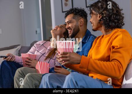 Verschiedene Freunde lachen, teilen sich gestreifte Popcorn-Wannen und halten die Fernbedienung auf der Couch im Wohnzimmer Stockfoto