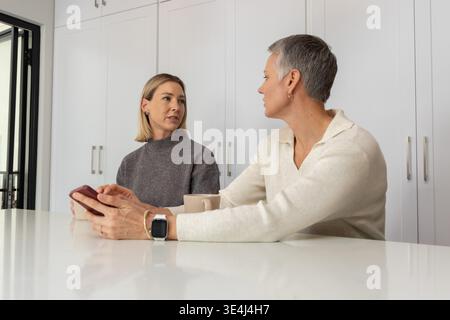 Ältere Frau und Frau mittleren Alters, die sich auf der Küchenarbeitsplatte beugen, Smartphone und Smartwatch halten Stockfoto