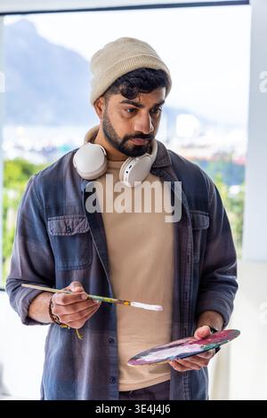 Männlicher Maler, der im Atelier steht und Farbe auf Palette und Pinsel vorbereitet, mit Mütze, Kopfhörern Stockfoto