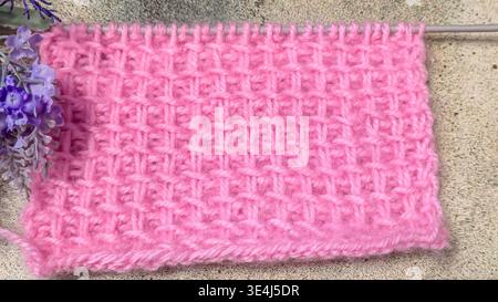 Ein strukturiertes, durchbrochenes Strickmuster aus farbigem Garn auf einer Holzoberfläche mit dekorativen Blumen. Nahaufnahme der Strickstruktur und des Strickmusters Stockfoto