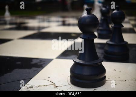 Eine Nahaufnahme eines großen schwarzen Plastikspielers, der auf einem feuchten, abwechselnden Schachbrett aus schwarzem und weißem Stein steht. Der Bauer ist in scharf f Stockfoto