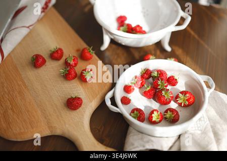 Frische Bio-Erdbeeren auf Holzbrett und Vintage-Keramikschalen. Trendige rustikale Küche mit roten Beeren, weißem Sieb und Sonnenlicht Stockfoto
