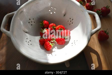 Frische rote Erdbeeren in Vintage-Keramiksieb auf Holztisch. Bio-Beeren in einer Schüssel aus weißem Steingut mit Sonnenlicht-Schatten, inspiriert von Wabi Stockfoto