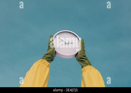 Ein Paar grüne Handschuhe halten eine weiße Uhr vor einem klaren blauen Himmel. Die Person, die hellgelbe Ärmel trägt, hebt die Uhr hoch und transportiert die Stockfoto