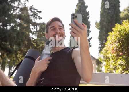 Ein lächelnder junger Mann macht ein Selfie nach einem Yoga-Workout im Freien, hält eine Wasserflasche und eine Matte in einem sonnigen Park mit Bäumen und genießt Fitness und Wohlbefinden Stockfoto