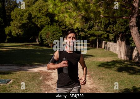 Sportlicher Mann, der im Freien in einem sonnigen Park trainiert, eine Wasserflasche hält und Sportausrüstung trägt. Rotes Muttermal am Hals sichtbar, fördert die Fitness. Stockfoto