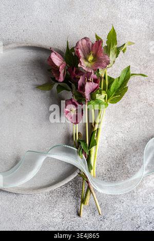 Minimalistischer flacher Lagen von purpurroten Blüten, gebunden mit einem weichen Band auf einem grauen Steinhintergrund, zarten Blütenblättern und grünen Blättern, natürliches B Stockfoto