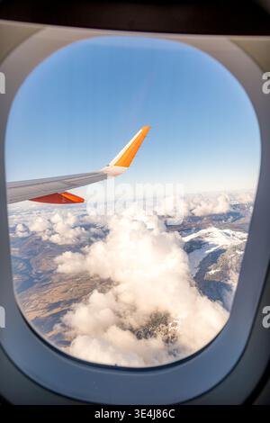 Ein atemberaubender Blick auf flauschige Wolken und schneebedeckte Berge durch ein Flugzeugfenster, wobei der Flügel des Flugzeugs sich in die klare Luft ausdehnt Stockfoto