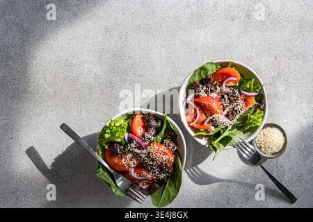 Blick von oben auf frische Spinat-Salatschalen mit Tomatenscheiben, schwarzen Oliven, roten Zwiebeln und Sesamsamen, serviert mit einem erfrischenden Apfelwein-Getränk auf einem gr Stockfoto