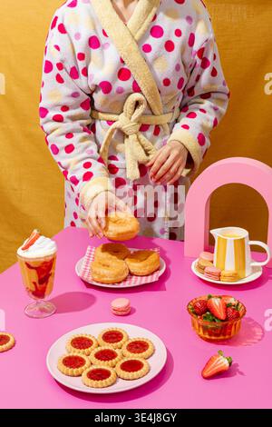 Eine Person in einem kuscheligen Bademantel arrangiert auf einem rosafarbenen Tisch mit Strohhalm ein Frühstück mit Donuts, mit Marmelade gefüllten Keksen und Makronen Stockfoto