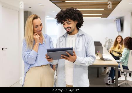 Ein lächelnder latin man zeigt einem Kollegen in einem hellen Coworking-Büro einen Tablet-Bildschirm, während Teamkollegen im Hintergrund arbeiten und Teamwork hervorheben Stockfoto