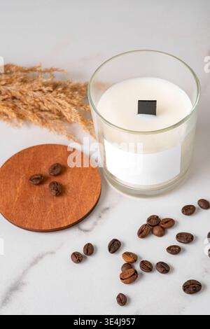 Minimales Stillleben mit einer weißen Kerze in einem Glasgefäß auf einer Marmoroberfläche, verstreuten Kaffeebohnen und einem hölzernen Untersetzer mit getrockneten Gräsern für A Stockfoto