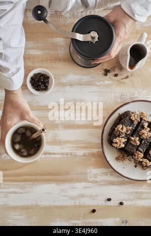 Blick von oben auf die Hände, die Kaffee mit einem manuellen Mahlwerk zubereiten, eine Tasse mit Eis, Bohnen und einem kleinen Krug auf einem rustikalen Tisch, neben einem Teller aus Schokoladenbraun Stockfoto