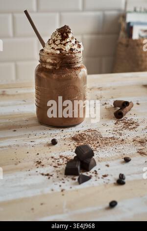 Rustikale Mokahfrappe, serviert in einem einmachglas mit Strohhalm, garniert mit Schlagsahne und Kakao. Schokoladenstücke, Kaffeebohnen und Zimtstangen drauf Stockfoto